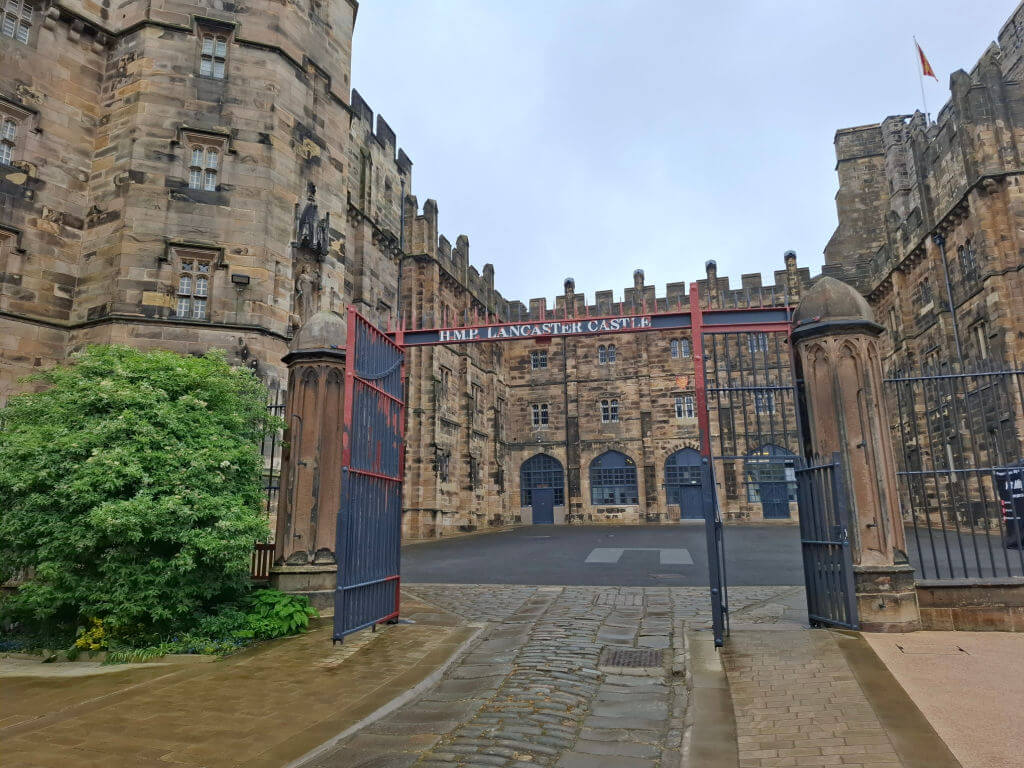 Historic Lancaster Castle with medieval stone walls and arched windows. An open, iron gate on a cobblestone path leads into the courtyard. A sombre atmosphere.
