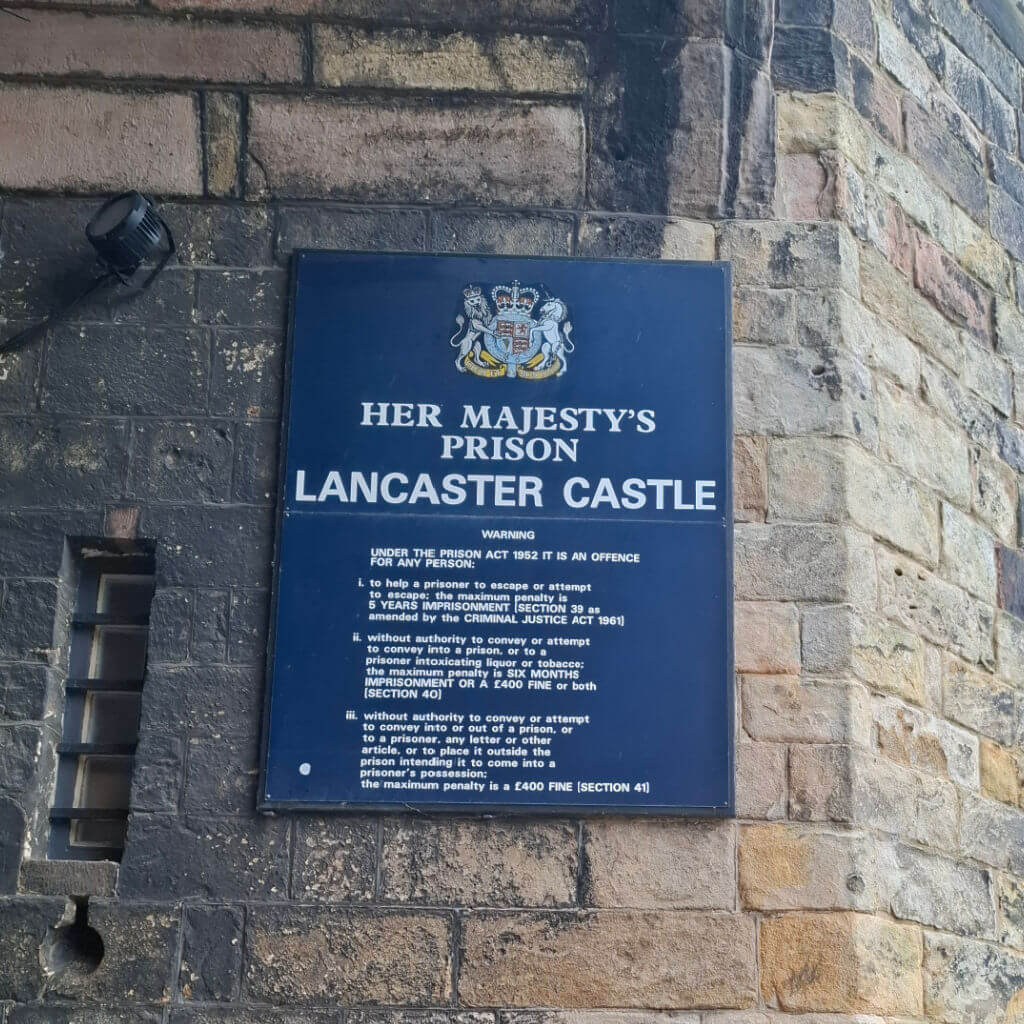 Blue sign on brick wall reads "Her Majesty’s Prison Lancaster Castle" with legal text and penalties. Old, historic tone with formal emblem above.