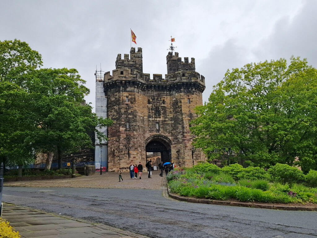 Old stone castle gate with two round towers, a flag flying atop. People with umbrellas walk on cobblestones. Lush green trees flank the path. Cloudy sky.