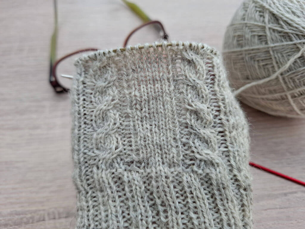 A close-up of a partially knitted sock with intricate cable patterns in light grey yarn. A pair of glasses and a ball of yarn rest on the wooden table.