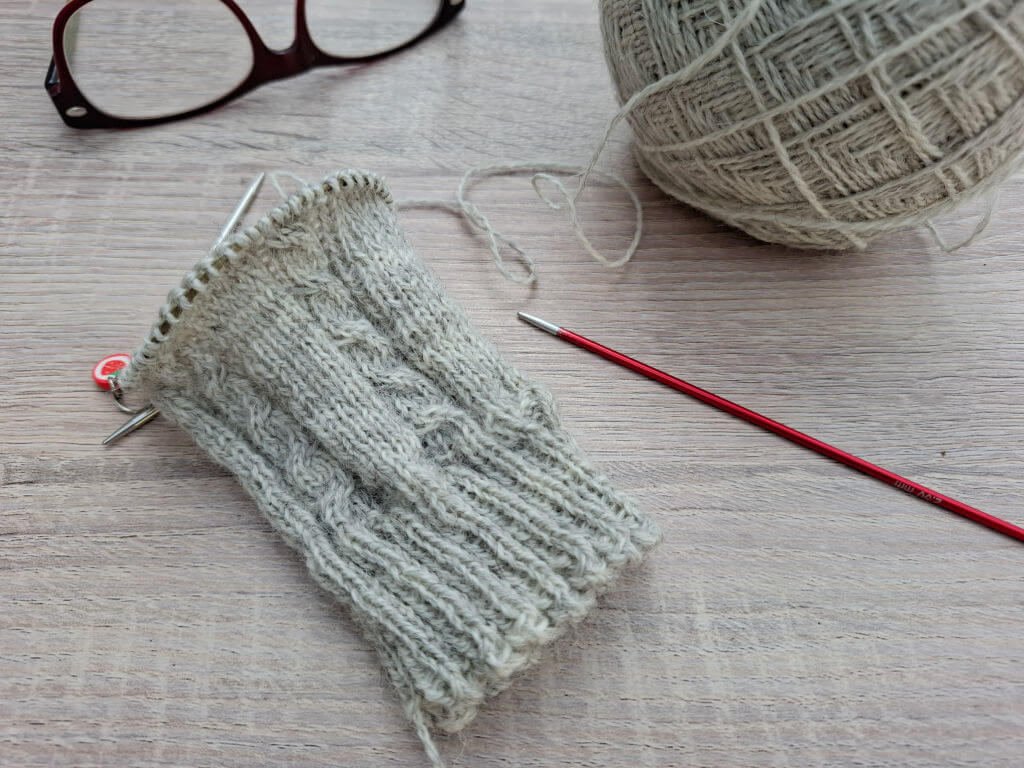 A partially knitted grey sock on needles lies on a wooden surface, with a ball of yarn and a pair of glasses nearby, conveying a cosy, creative vibe.