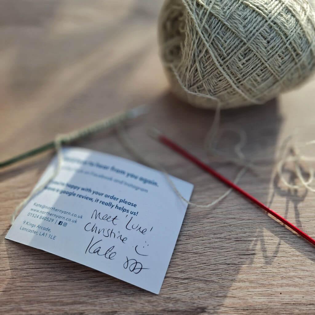 A ball of grey yarn, knitting needles, and a handwritten note on a wooden table. The note expresses thanks with a cheerful tone and smiley face.