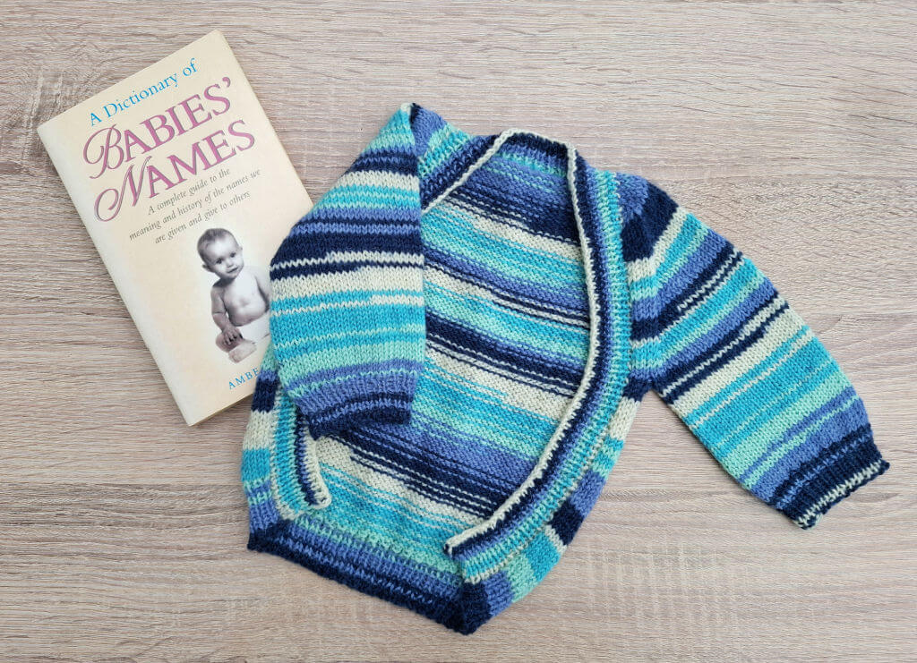 A colourful hand-knitted baby cardigan in blue, turquoise, and cream stripes next to a book titled "A Dictionary of Babies’ Names" on a wooden surface.