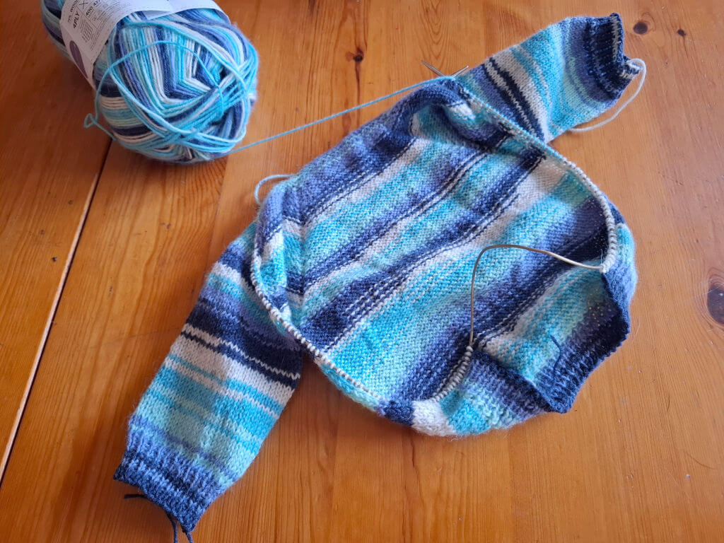 Knitted two-tone blue and white striped sweater in progress on wooden table, with matching yarn ball nearby, conveying a cosy, handmade vibe.