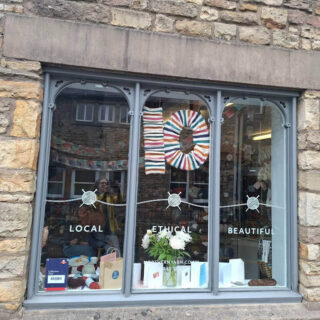 Shop window with "Local, Ethical, Beautiful" decals. Display features a colourful yarn "10" and white flowers, conveying a welcoming, creative vibe.