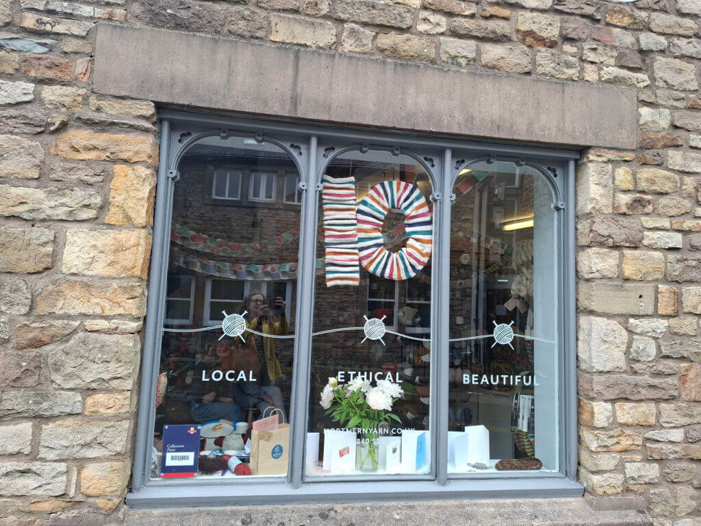 Shop window with "Local, Ethical, Beautiful" decals. Display features a colourful yarn "10" and white flowers, conveying a welcoming, creative vibe.