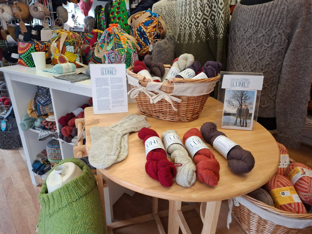 Cosy yarn shop display with colourful skeins, knitting patterns, and wool sweaters. A basket holds yarn, surrounded by vibrant fabric bags. Inviting and crafty.