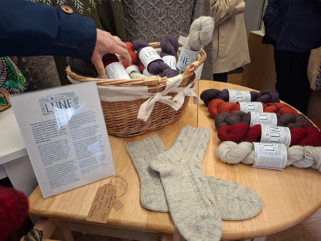 A display of colourful yarns in a wicker basket on a wooden table, accompanied by knitted socks and a sign. A hand reaches for a yarn skein.