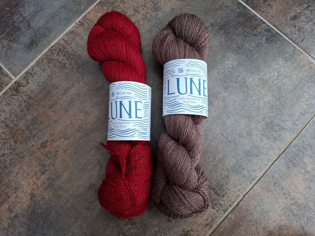 Two skeins of yarn are displayed on a tiled surface. The left skein is red, and the right skein is brown. Both have labels reading "LUNE."