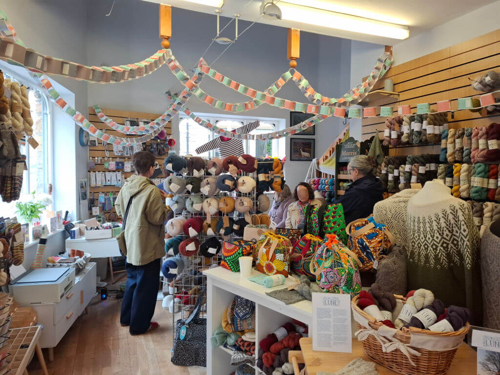 A cozy yarn shop filled with colourful skeins, knitted garments, and patterned bags. Two people browse, and festive paper garlands hang overhead.