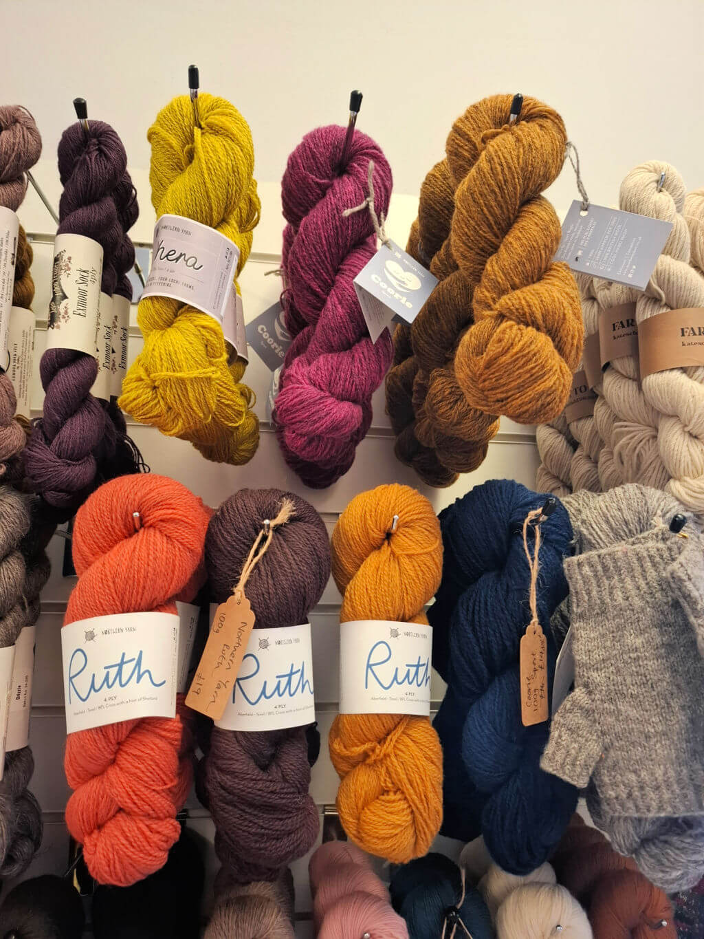 A display of colourful yarn skeins arranged on a white wall. Vibrant shades include mustard, magenta, orange, and blue, exuding a cosy, creative vibe.