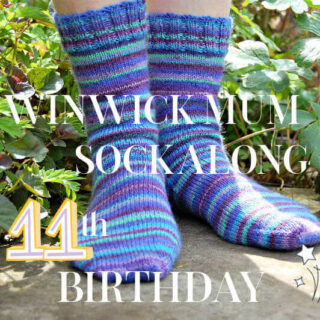 Two feet wearing colourful striped knitted socks in a garden setting with green foliage. Text overlay reads "Winwick Mum Sockalong 11th Birthday."