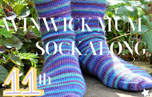 Two feet wearing colourful striped knitted socks in a garden setting with green foliage. Text overlay reads "Winwick Mum Sockalong 11th Birthday."