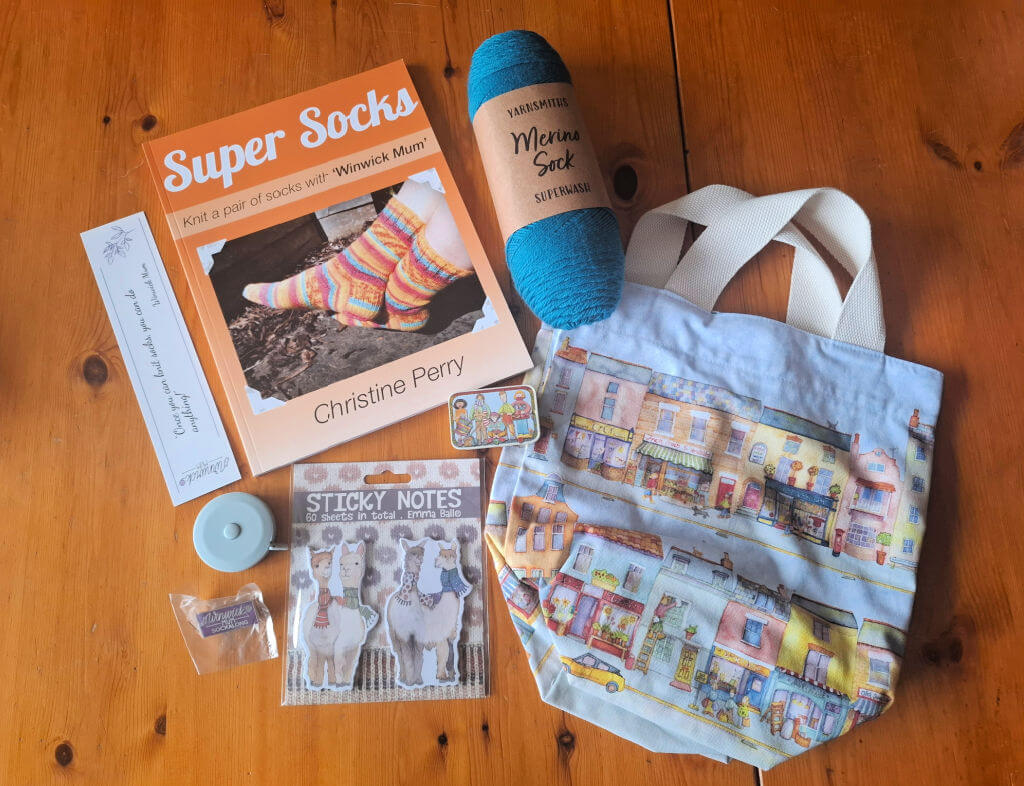 A knitting-themed flat lay on a wooden surface, featuring a "Super Socks" book, blue yarn, illustrated tote, llama sticky notes, tape measure, bookmark, and small tin.