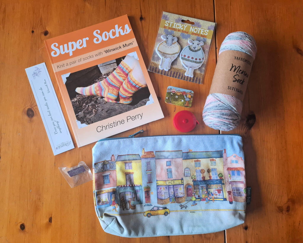 Knitting supplies on a wooden table, including a "Super Socks" book, yarn, sticky notes, tape measure, and a pouch with colourful buildings. Cosy and creative vibe.