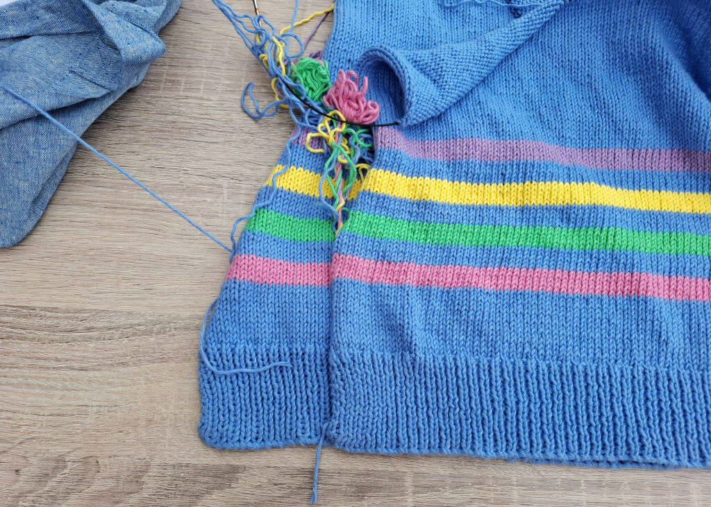 Close-up of a blue knitted sweater with unraveling multicoloured stripes in pink, green, and yellow. Loose yarn ends create a messy look.