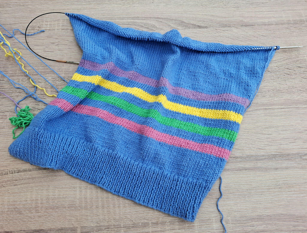 Knitting project with blue yarn featuring horizontal stripes in pink, green, yellow, and purple. Incomplete with needles and loose threads visible.