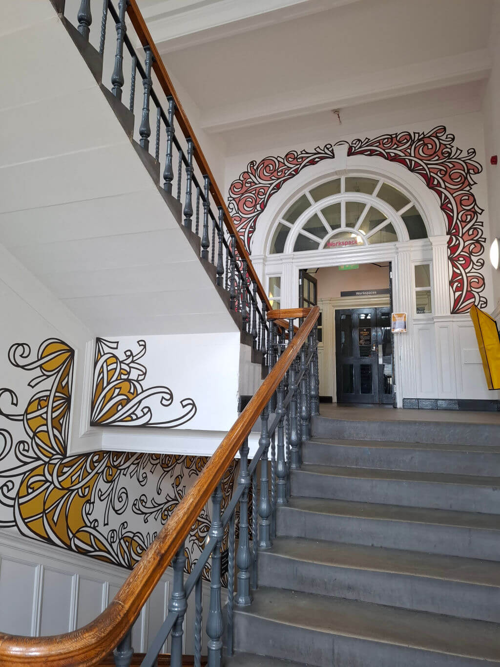 Historic staircase with intricate black and yellow floral wall designs. Curved wooden banister, arched doorway, evokes artistic elegance.