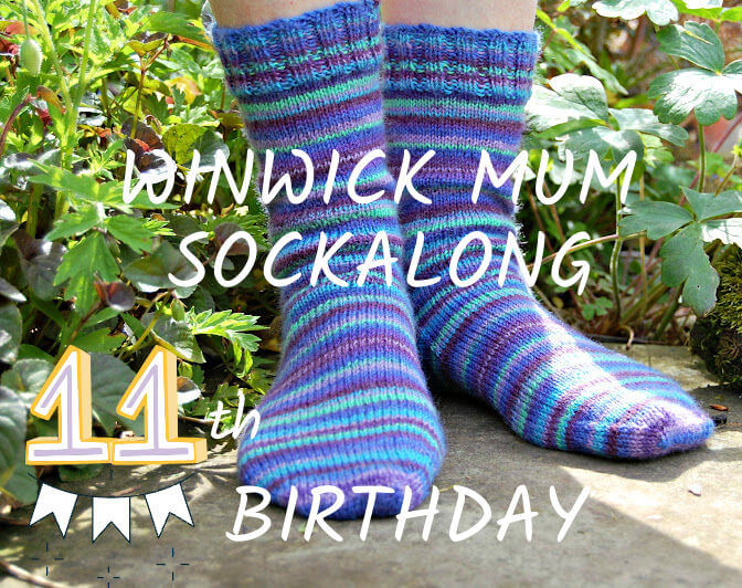 Two feet wearing colourful striped knitted socks in a garden setting with green foliage. Text overlay reads "Winwick Mum Sockalong 11th Birthday."