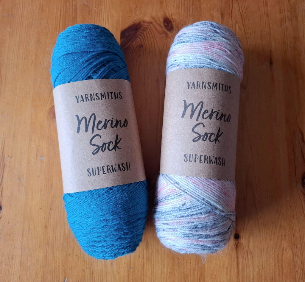 Two skeins of Yarnsmiths Merino Sock Superwash yarn on a wooden table. The left skein is teal, and the right is multicoloured in pastel shades.