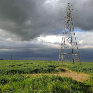 A tall metal electricity pylon stands in a green field under a dramatic sky. Dark storm clouds loom, casting a moody and tense atmosphere over the scene.