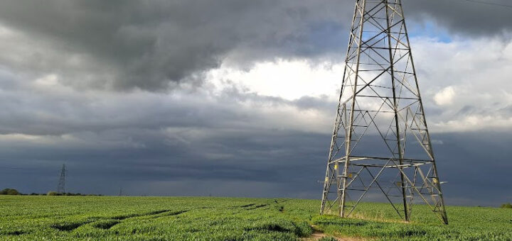 A tall metal electricity pylon stands in a green field under a dramatic sky. Dark storm clouds loom, casting a moody and tense atmosphere over the scene.