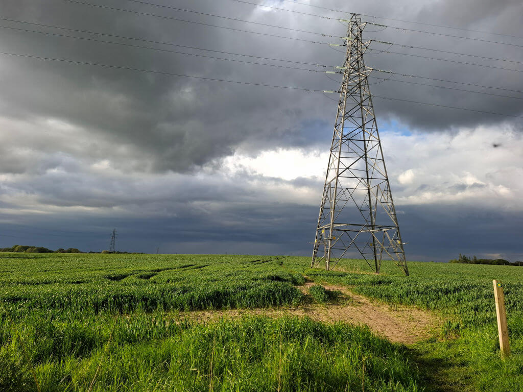 A tall metal electricity pylon stands in a green field under a dramatic sky. Dark storm clouds loom, casting a moody and tense atmosphere over the scene.