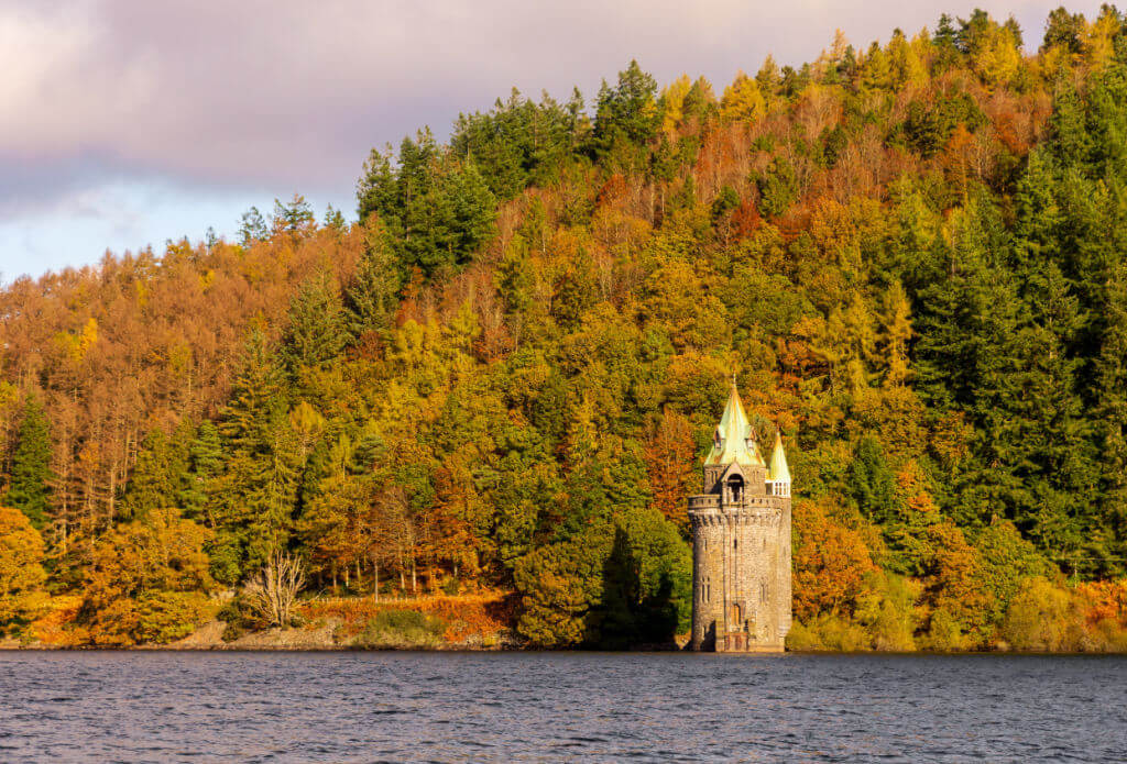 A solitary stone tower with pointed roofs stands by a tranquil lake, surrounded by a densely wooded hillside in vibrant autumn colours under a cloudy sky.