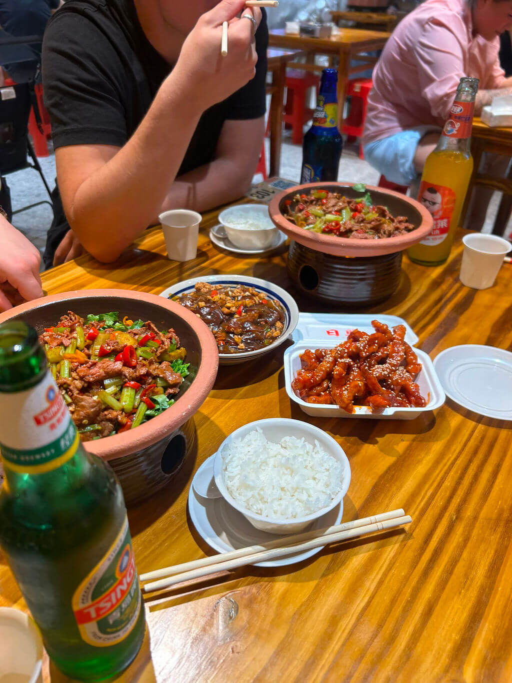 A table with Asian dishes includes clay pots of stir-fry, a plate of glazed meat, rice bowl, and beer bottles, with people dining, creating a lively atmosphere.
