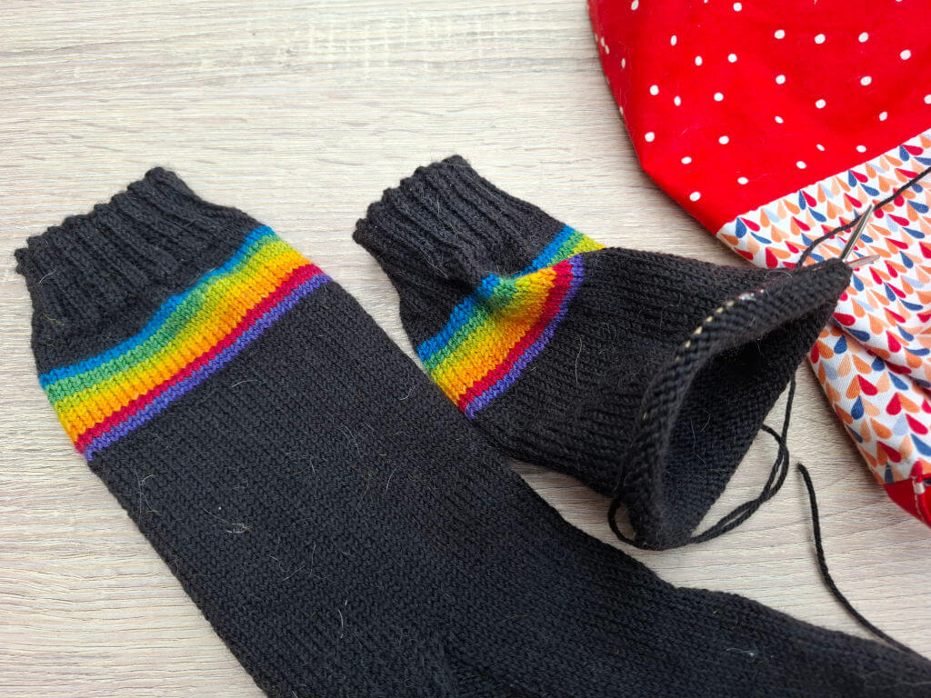 Black socks with vibrant rainbow stripes at the cuffs, lying on a wooden surface. A red polka dot and patterned bag is partially visible.