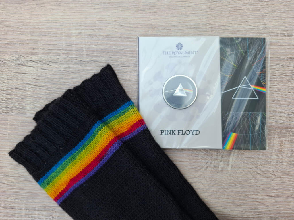 Black socks with rainbow stripes lie next to a Pink Floyd-themed Royal Mint coin in packaging. The coin features a prism design.