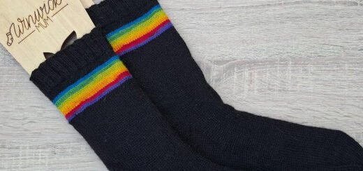 Black socks with vibrant rainbow stripes near the top, displayed with wooden sock blockers. The scene conveys warmth and comfort.