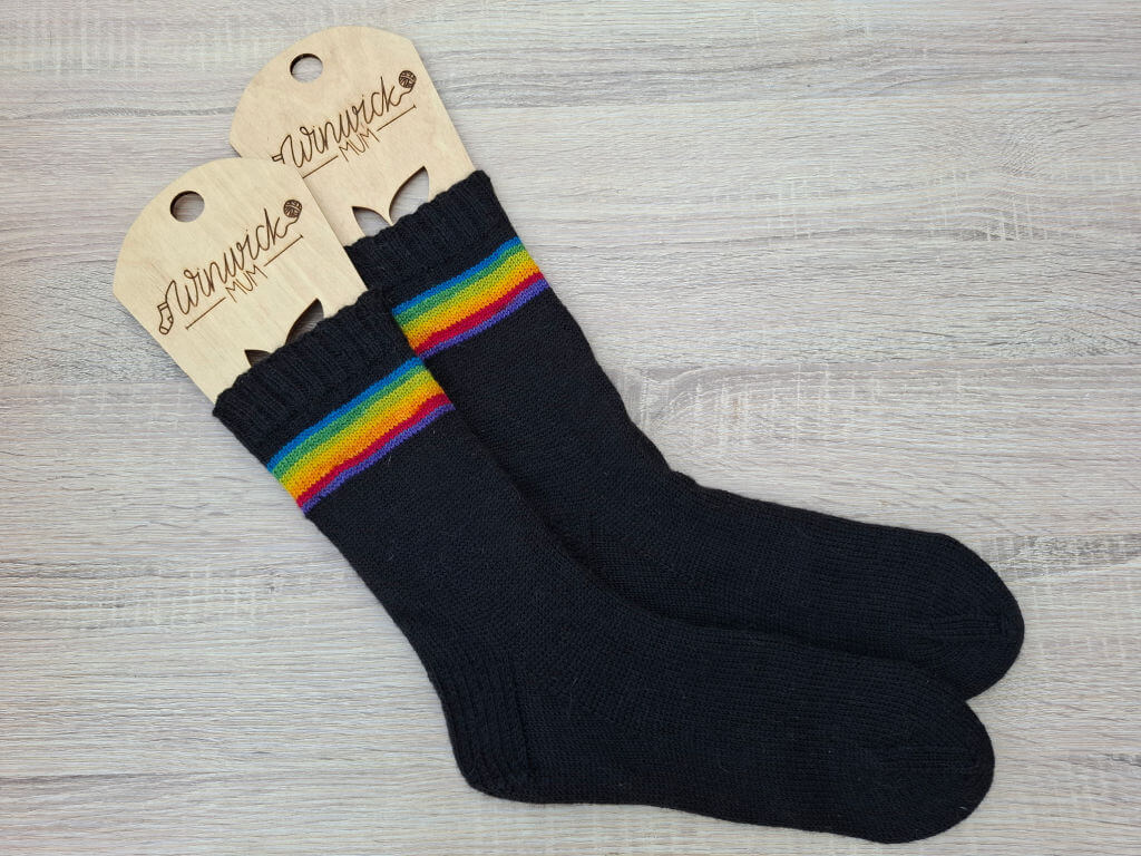 Black socks with vibrant rainbow stripes near the top, displayed with wooden sock blockers. The scene conveys warmth and comfort.