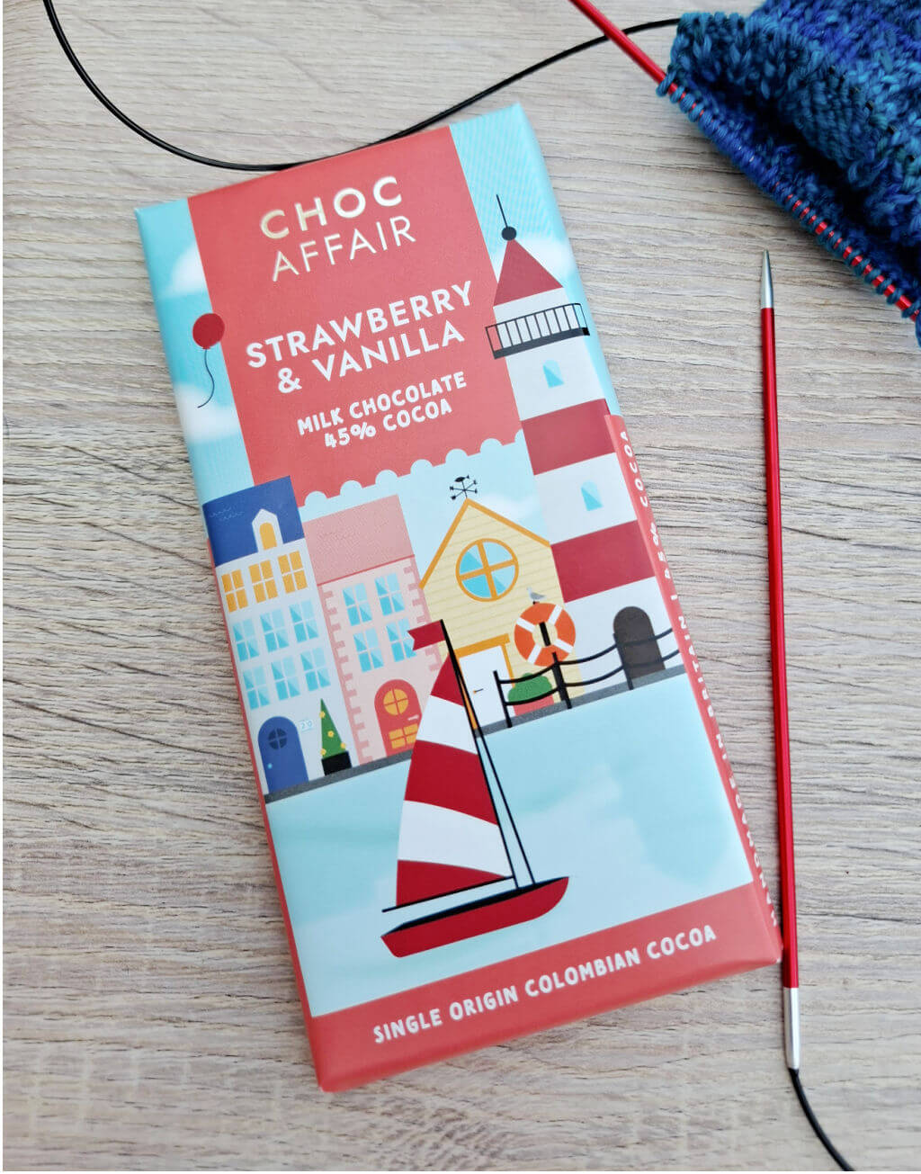 Colourful chocolate bar packaging on a wooden surface. It features a sailing boat and houses with a knitting project and needle nearby. The label reads 'Choc Affair, Strawberry & Vanilla.' The scene conveys a cosy, creative vibe.