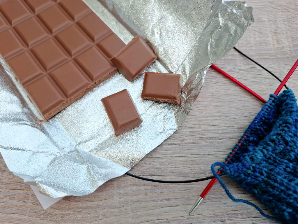 An unwrapped chocolate bar with some squares broken off lies beside blue knitting on red needles, resting on a light wooden surface.