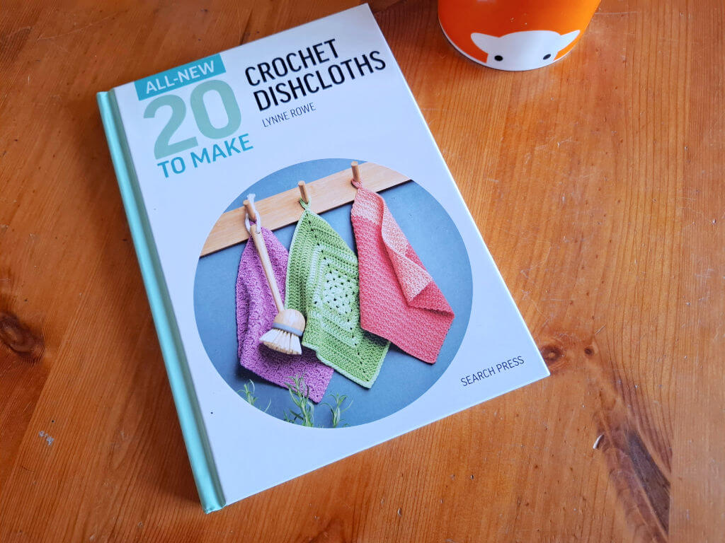 A book titled "20 to Make: Crochet Dishcloths" lies on a wooden table. The cover shows three colourful crocheted dishcloths hanging on pegs.