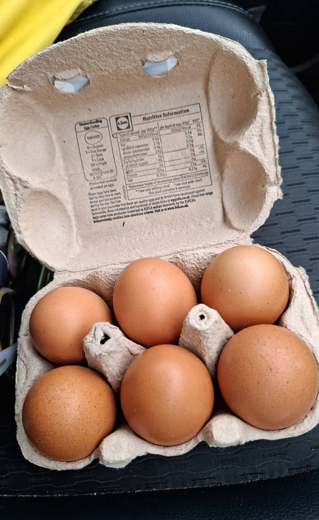 A cardboard egg carton with six brown eggs is open, showing nutrition information inside the lid. The carton rests on a textured black surface.