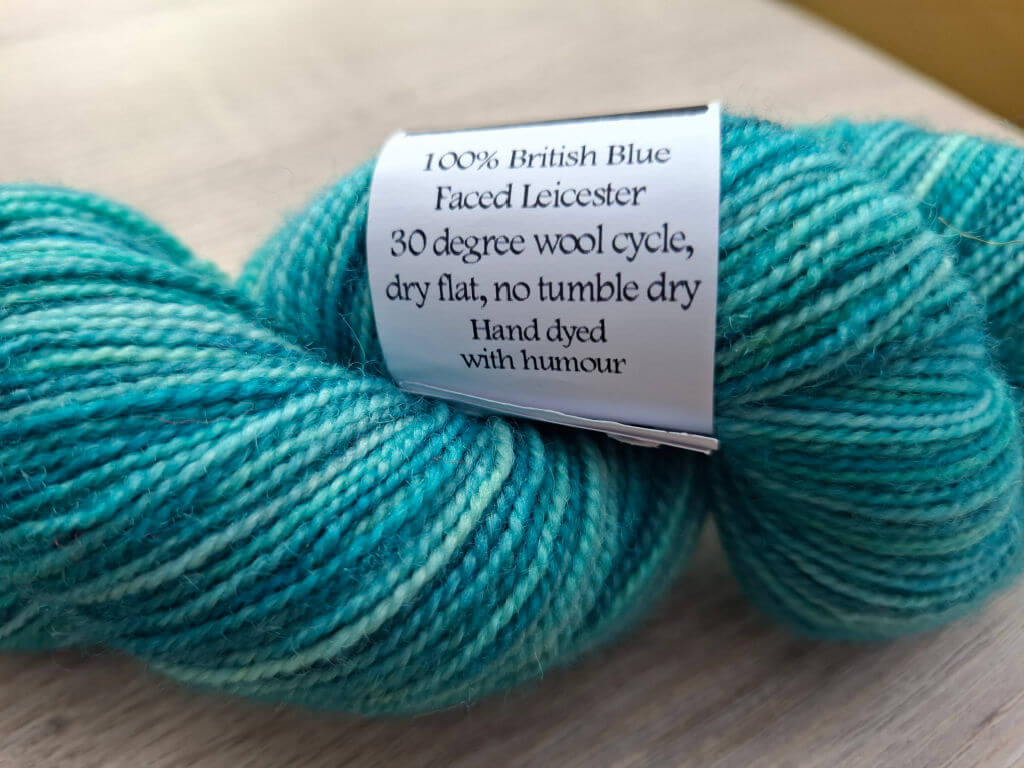 A skein of teal yarn labeled "100% British Blue Faced Leicester." Care instructions are "30 degree wool cycle, dry flat, no tumble dry." Text adds humor.