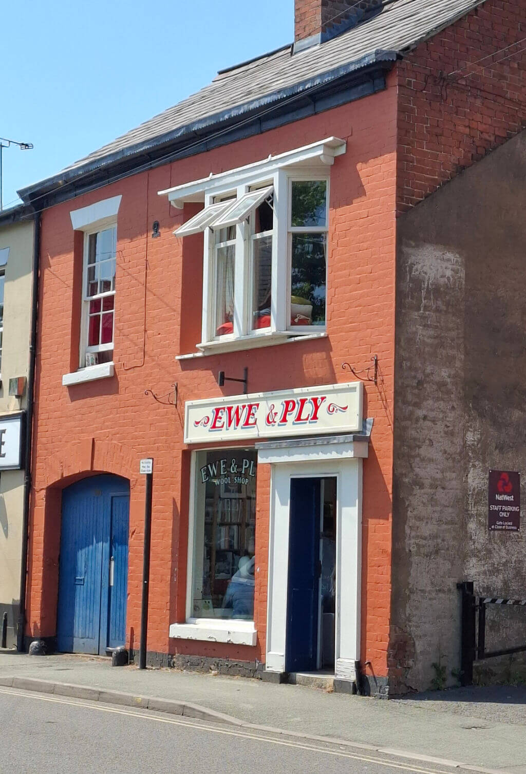 A charming yarn shop, "Ewe & Ply," in a quaint brick building with an painted facade. Blue door, large windows, and a cosy, inviting atmosphere.