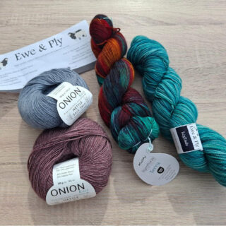Four skeins of yarn sit on a wooden table: blue, burgundy, multicoloured, and teal. A paper labeled "Ewe & Ply" lies partially beneath the blue yarn.
