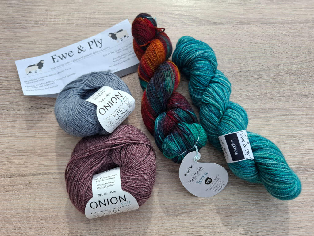 Four skeins of yarn sit on a wooden table: blue, burgundy, multicoloured, and teal. A paper labeled "Ewe & Ply" lies partially beneath the blue yarn.
