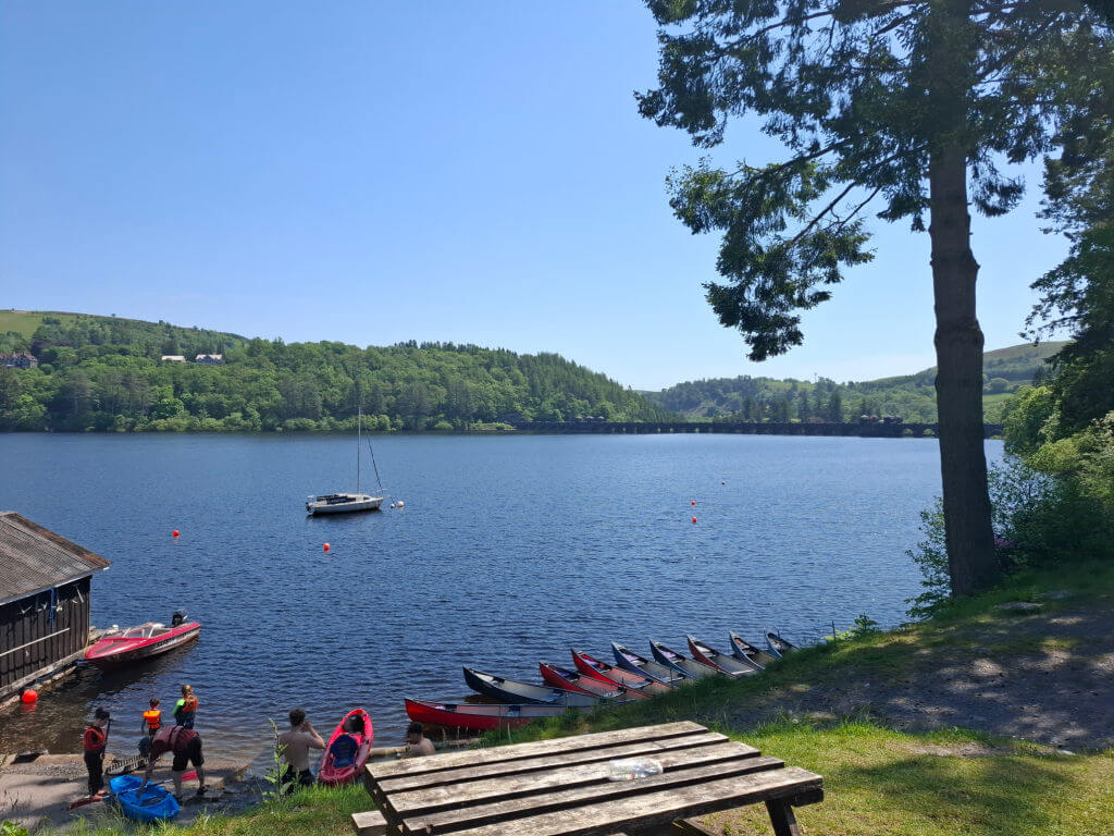 A serene lake with dark blue water, surrounded by lush green trees under a clear blue sky. Canoes line the shore, and a lone sailboat is on the water.