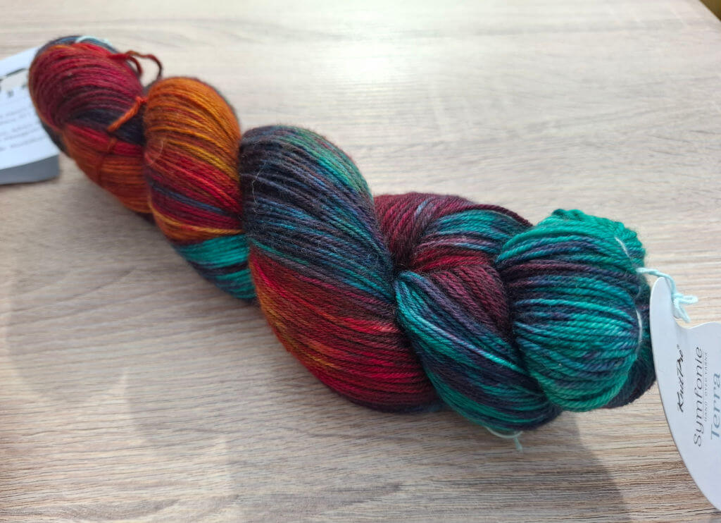 A skein of multicoloured yarn rests on a wooden surface, displaying rich hues of red, orange, teal, and purple. A tag is attached at one end.