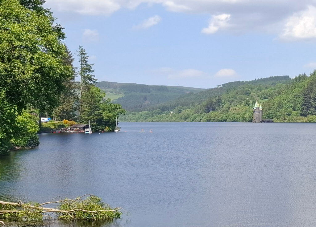 A serene lake scene with trees lining the shore, a distant stone tower, and lush hills under a bright blue sky. The mood is peaceful and scenic.