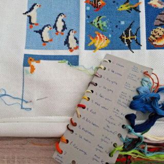 Cross-stitch artwork in progress features penguins and tropical fish. Threads and a colour guide are visible on the wooden frame.