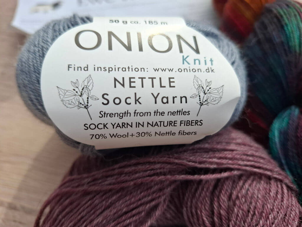 Close-up of a ball of Onion Knit Nettle Sock Yarn. The label reads "Strength from the nettles, 70% Wool + 30% Nettle fibers." Soft, earthy tones.