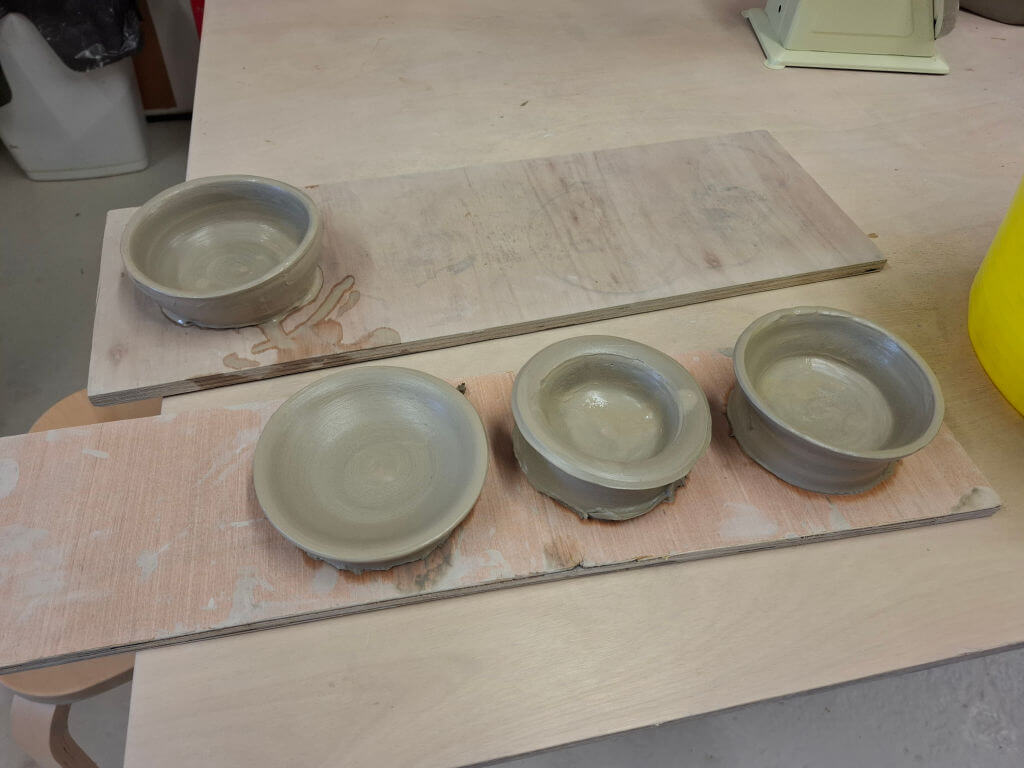 Four unglazed clay bowls sit on wooden boards in a pottery studio, reflecting a sense of calm and craftsmanship. A bright yellow bucket is partially visible.