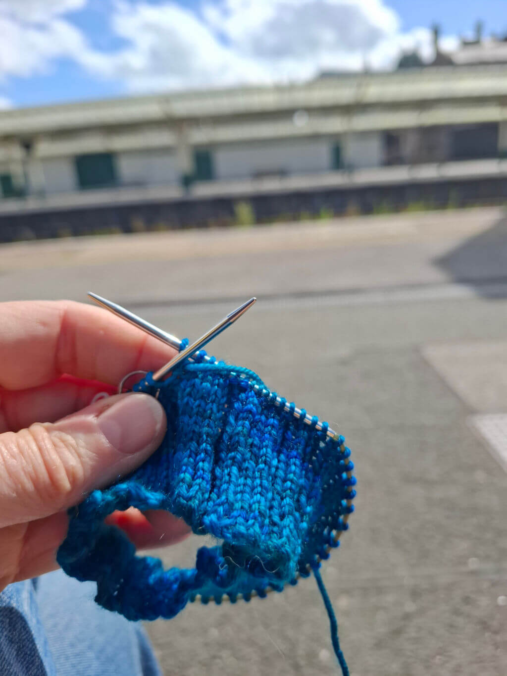 A hand holding knitting needles and blue yarn, with a partially knitted sock. The background shows a sunny day at an empty train station platform.