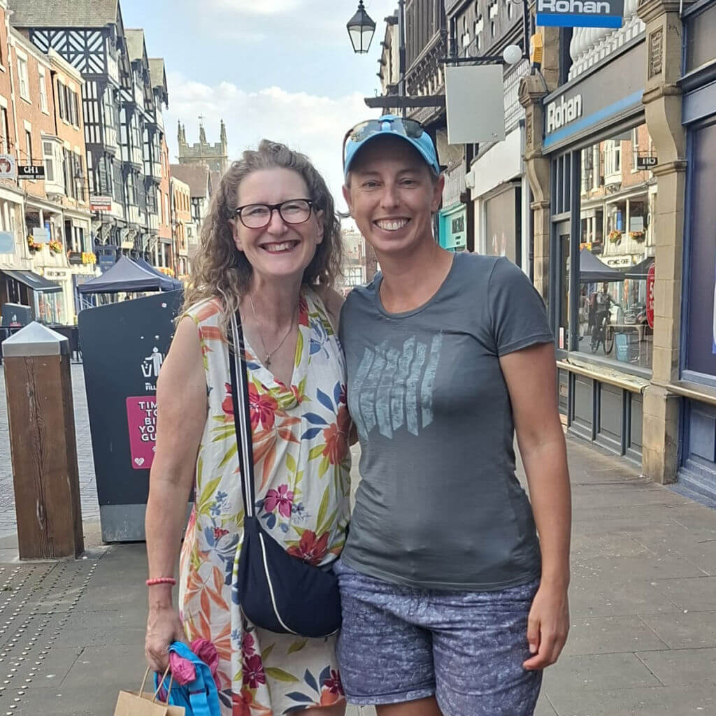 Two women smile at the camera.  They are standing in a street with historic black and white buildings.
