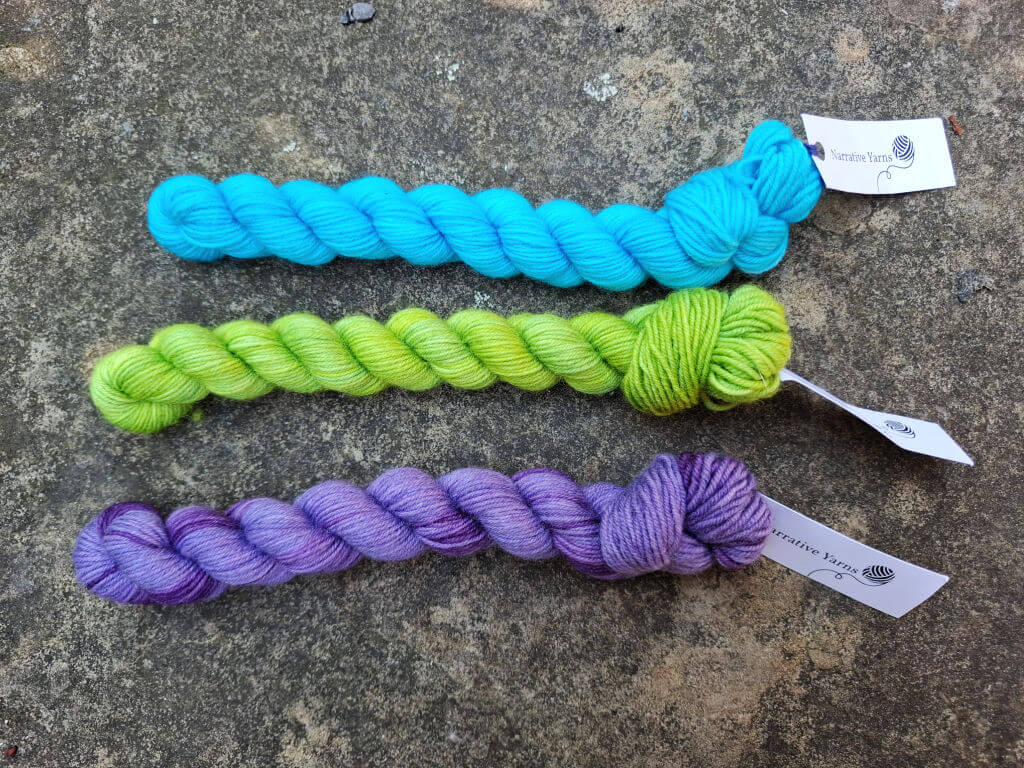 Three twisted skeins of yarn lie on a textured gray surface. From top to bottom, they are turquoise, lime green, and purple, each with a small tag.